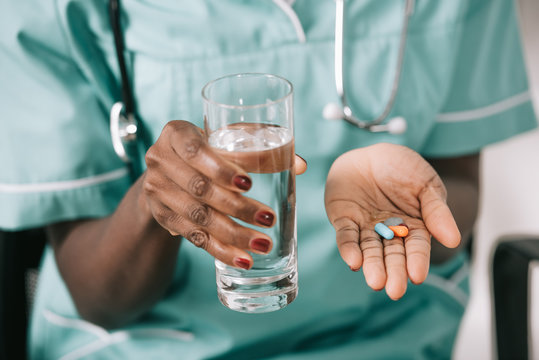 cropped view of african american nurse with stethoscope holding glass of water and pills in hand