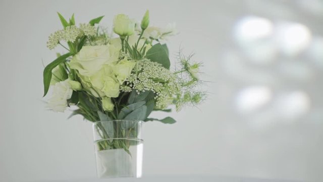 Camera shows nice bouquet of white flowers with big green leaves in glass vase put on white table in room with grey walls.