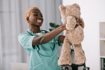 smiling african american nurse with stethoscope holding teddy bear