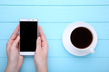Female hands holding smartphone and cup of coffee on blue wooden background.
