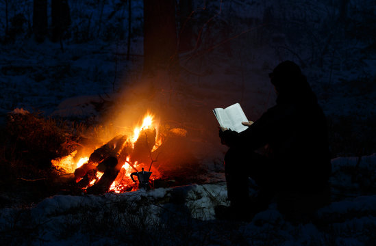 Man Reading A Book By The Fire. Book Is Highlighted By Headlamp Winter Night In The Forest