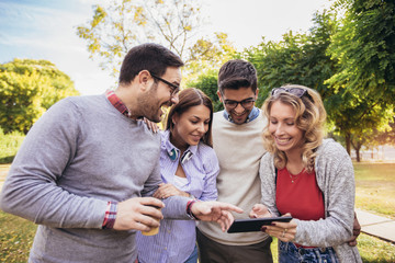 Four happy smiling young friends walking outdoors in the park holding digital tablet