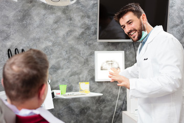 Smiling young male dentist showing x-ray images to a patient, consulting during the exam in dental...