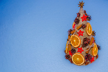 Необычная новогодняя ель, выполненная в стиле «арт-деко» украшенная сушеными апельсинами, сосновыми шишками, бадьяном, корицей и расположенной на снежном покрове. - obrazy, fototapety, plakaty