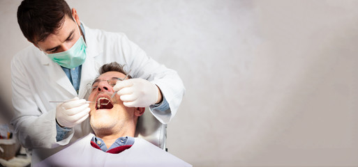 Young male dentist holding a tooth mirror and dental pick, performing dental exam on a mature male...