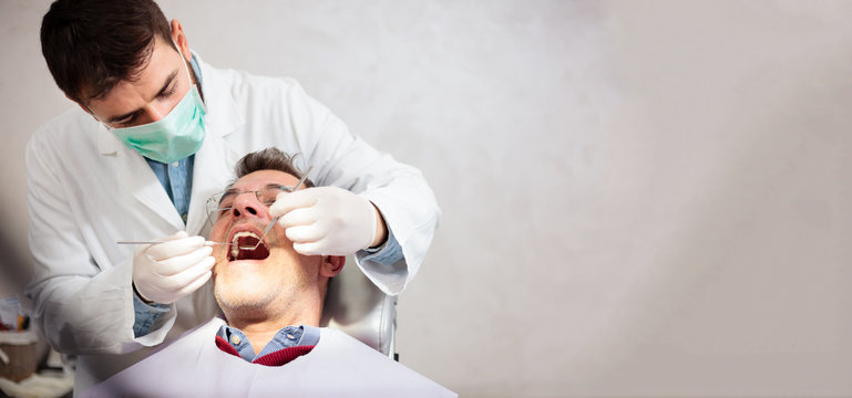 Young male dentist holding a tooth mirror and dental pick, performing dental exam on a mature male patient in dental clinic. Wide ratio image with copy space on the right side.