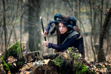 Beautiful young woman warrior with blue eyes and specific makeup wearing ram horns and fur collar hiding behind stumps in forest, stalking and ready to attack with ax.
