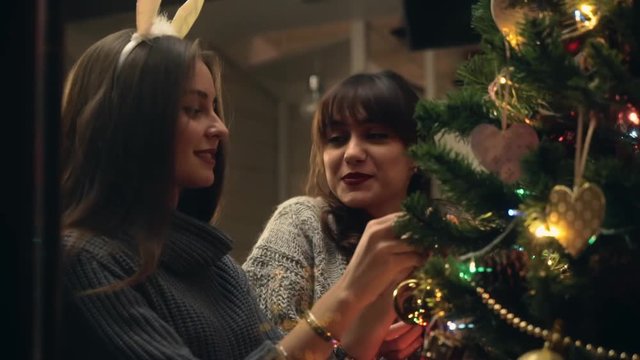 Girlfriends decorate a christmas tree with joy and in festive mood. Two girls Laughing while dressing up christmas tree. Sisters together fill the Christmas tree with toys, preparing for holidays