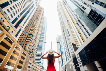 Young pretty woman in red dress and straw hat walking beetween skycrapers in modern city. Low angle view