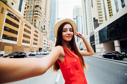 Young pretty woman in red dress and straw hat take selfie photo in front of skycrapers of modern arhitecture city.