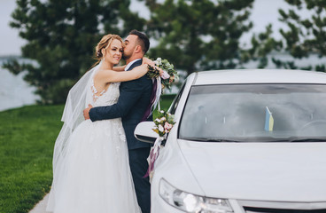 Stylish newlyweds are hugging near a white car in a green park, against the backdrop of summer nature. Bearded groom and blond bride in a lace dress are smiling outdoors. Wedding portrait.