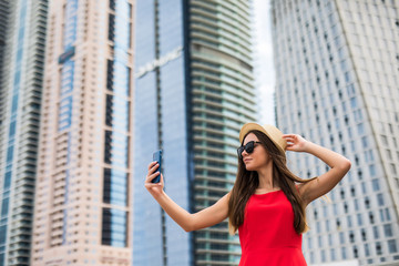 Portrait of young smile woman in red dress, sunglasses and summer hat take selfie on the phone on downtown skycrapers background