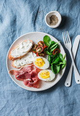 Cream cheese toast, boiled egg, prosciutto, spinach, tomatoes - delicious healthy breakfast or snack on blue background, top view