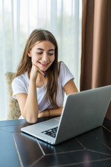 Fototapeta na wymiar Young smiling girl is sitting on modern chair near the window in light cozy room at home working on laptop in relaxing atmosphere
