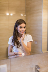 Naklejka premium Beautiful young woman cleaning her skin with a cotton pad, looking at the mirror at home bathroom. Beauty, skin care concept, lifestyle