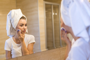 Beautiful young woman is treating her face after shower standing and touching her cheek with a sponge. The woman is holding a mirror and smiling