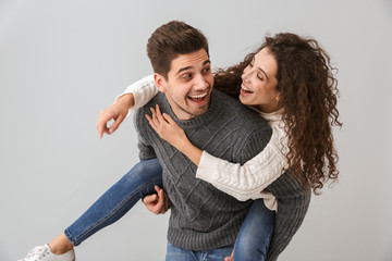 Image of joyful couple having fun and while man piggybacking attractive woman, isolated over gray...