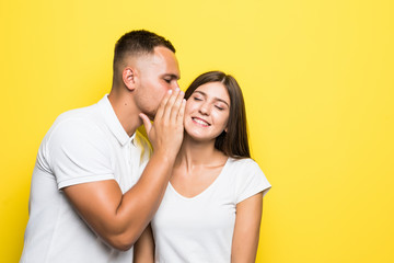 Portrait of handsome man whispering secret or interesting gossip to young woman in her ear isolated over yellow background