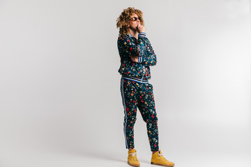 Adult shylish man in fashionable vintage style tracksuit and sneakers posing on white background. Funky man stroking  beard. Awesome confident male in sunglasses portrait. 80s fashion. Serious person.