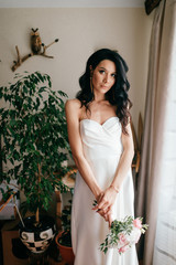 Pretty young caucasian bride with bouquet of flowers posing indoor. Lovely girl with spanish appearance closeup toned  home portrait. Dark eyed bride in white dress with pensive face tender fellings.