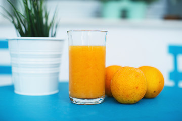 A fresh orange juice with some oranges nearby placed on a blue table with a table plant