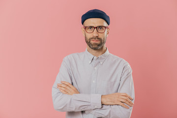 Self confident unshaven man crosses hands over chest, wears spectacles and white shirt, isolated...
