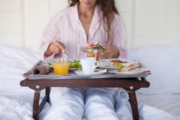Obraz na płótnie Canvas Making breakfast in bed / pretty smiling brunette woman holding a tray with healthy food while sitting on the bed