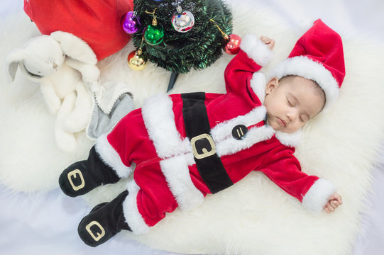 little baby wearing Santa Claus costume sleep on white fur carpet with Christmas tree. Concept of celebrates Christmas and New Year's holidays.