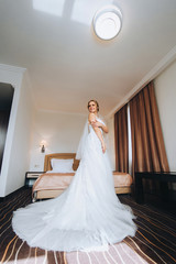 Fototapeta premium A lovely bride with a long dress is standing in a beautiful room and smiling. Wedding interior. Charges the bride at the hotel. Wedding photography. Portrait of a beautiful bride.