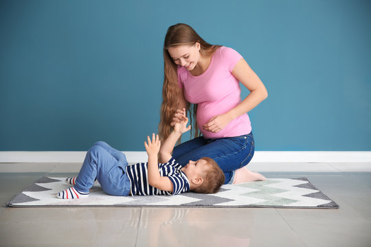 Young pregnant woman playing with her cute little son on carpet near color wall