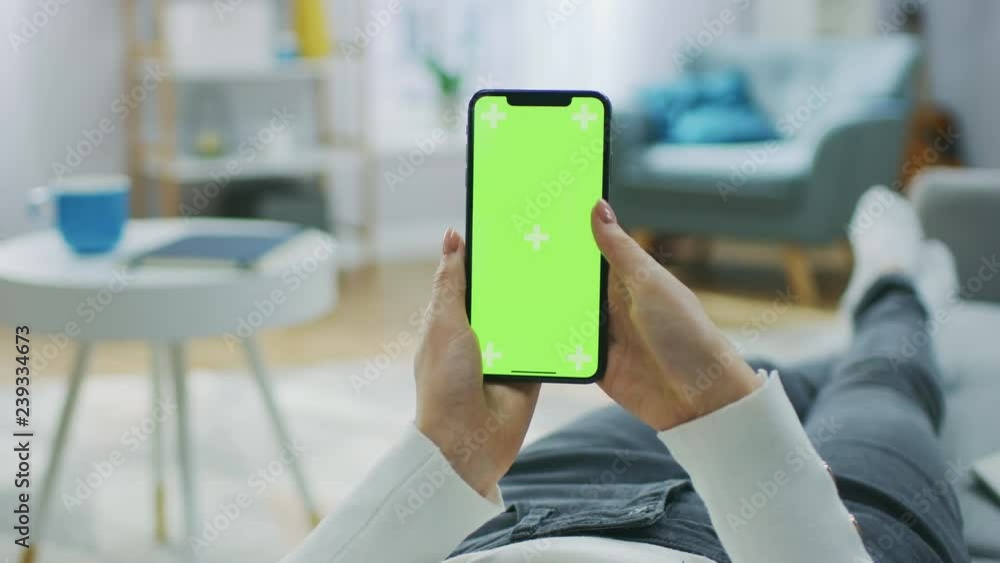 Poster woman at home lying on a couch using smartphone with green mock-up screen, doing swiping, scrolling 