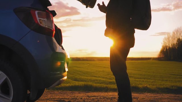 Tourist is opening car trunk in the middle of field in golden sunlight. Trunk is opening by athletic male and he puts backpack inside the car