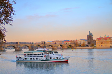 Naklejka premium Pleasure boat on the Vltava River in Prague against the backdrop of Charles Bridge at sunset.