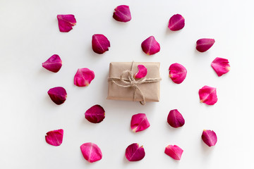 Gift in craft paper with pink rose petals background on white table. Romance concept. Flat lay, top view, copy space