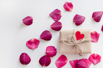Gift in craft paper with red heart decoration on pink rose petals background on white table. Romance concept. Flat lay, top view, copy space