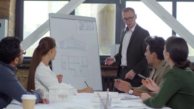 Young male architect in eyeglasses and formal suit drawing building construction plan on flipchart and explaining it to interracial team of colleagues while giving presentation during office meeting