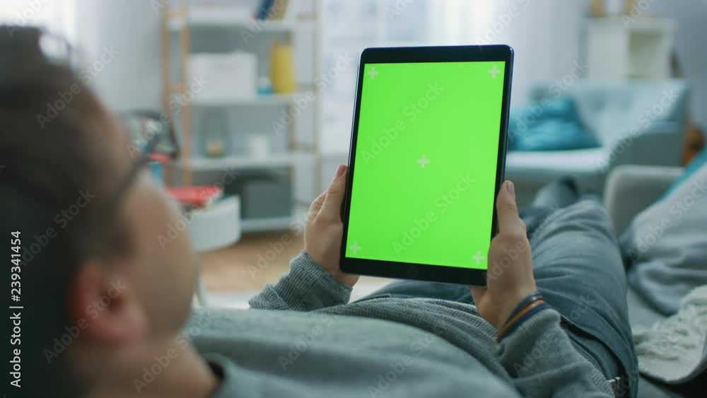 Poster young man at home laying on a couch using with green mock-up screen tablet computer in vertical port