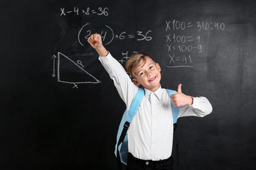 Cute little schoolboy with backpack showing thumb-up near blackboard