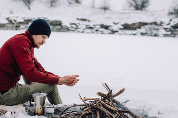 Obraz premium tourist is sitting near camp fire an in winter time, surrounded by snow near of the frozen lake. Concept adventure active vacations outdoor. Winter camping