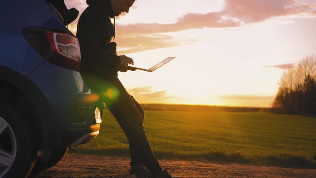 A tourist stands near the car in the middle of a green field at sunset, holds in his hand a laptop looking at a map of the trip. He closes the laptop, puts it in the trunk and gets into the car