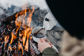 Traveling woman sits near camp fire an in winter time and pours itself hot coffee . Concept...