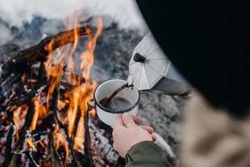 Fotobehang Reizende vrouw zit in de winter bij het kampvuur en schenkt zichzelf hete koffie in. Concept avontuur actieve vakanties buiten. Winterkamperen © Yura Yarema