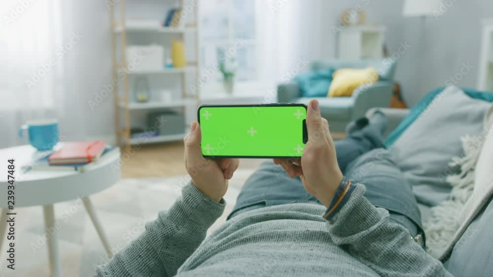 Poster man at home lying on a couch using smartphone, holds it horizontally in landscape mode. playing vide