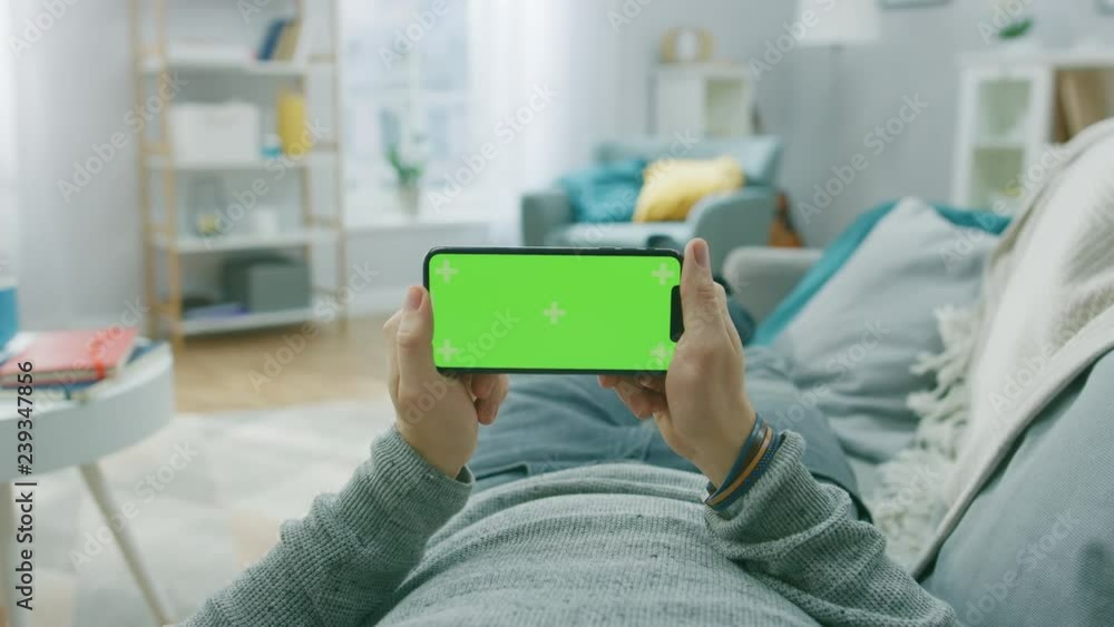 Poster man at home lying on a couch using smartphone, holds it horizontally in landscape mode. playing vide