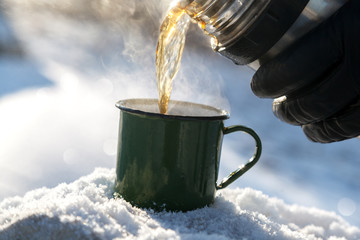 Pouring tea from a thermos in the open air. Cold snow and frosty air. Mug with hot tea and hot...