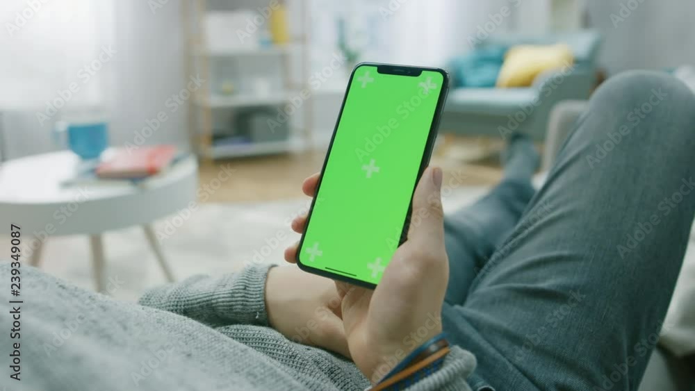 Poster man at home lying on a couch using smartphone with green mock-up screen, doing swiping, scrolling ge