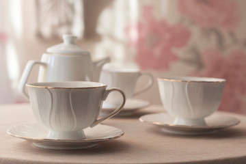 Obraz na płótnie Canvas Tableware for tea party - teapot and cups - on white tablecloth on light pink floral background with sunlight. Close-up, copy space