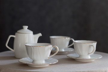 Tableware for tea party - teapot and cups - on white tablecloth on dark gray background. Close-up, copy space
