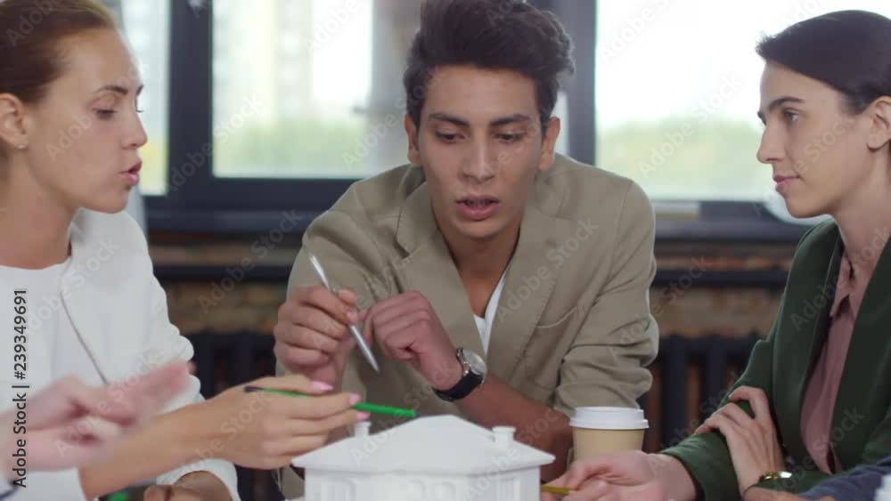 Poster young male architect pointing at paper model of residential building and discussing it with colleagu