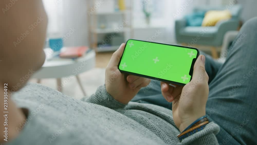 Poster man at home lying on a couch using smartphone, holds it horizontally in landscape mode. playing vide